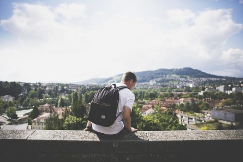 A male student wearing a backpack sits on a ledge overlooking a city.