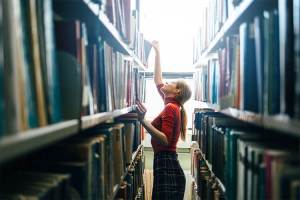 A young, female college student looking for a book on the shelves at her school library.