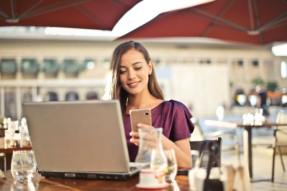 A smiling woman in a purple blouse, in an outdoor cafe, checks both her phone and laptop.