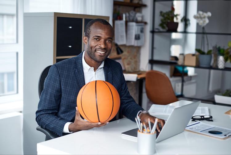 A sport business manager sits at an office desk while holding a basketball.