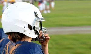 A profile shot of a young football player with a blue uniform and a white helmet. Other children can be seen on the grassy field in the background.