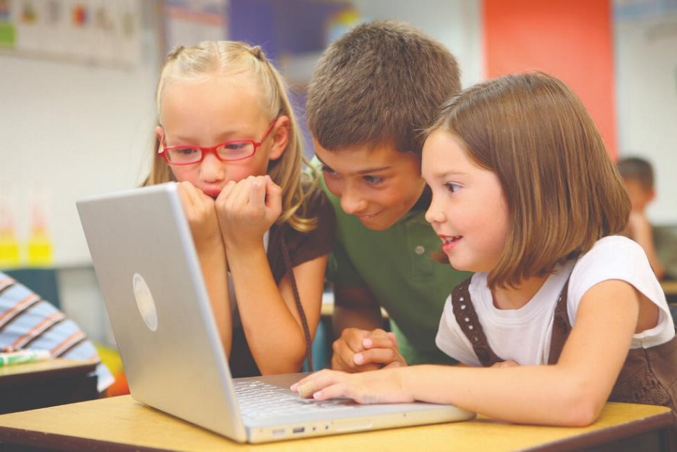 Three children surfing the web on a computer in a classroom monitored by a teacher