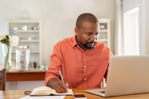 A man attending an online class on his laptop and taking notes.