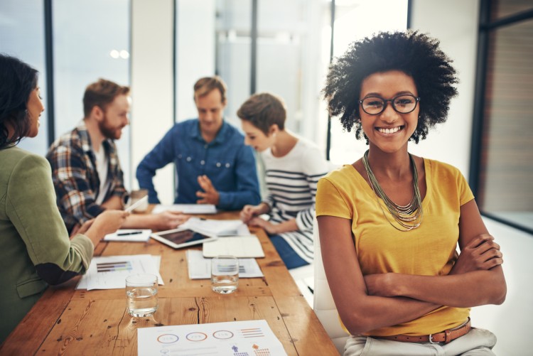 A smiling small business owner sits at a table with employees meeting behind them.