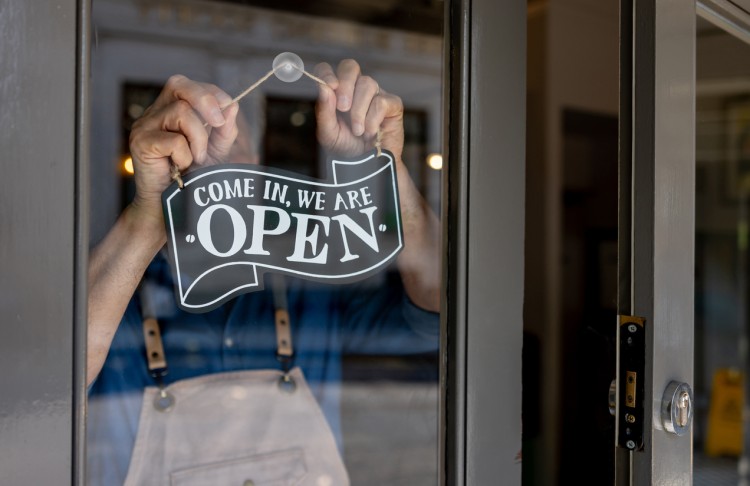 A small business owner hangs a sign on a glass door reading “Come in, we are open.”
