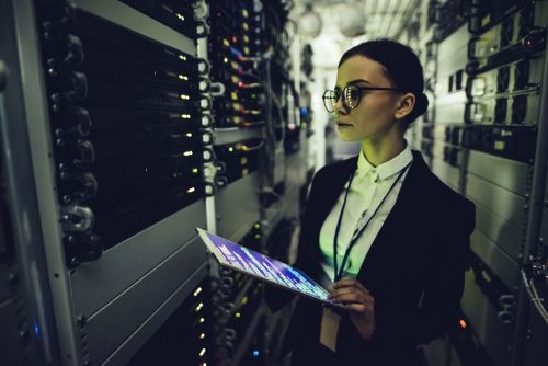 A woman with a tablet, taking advantage of her master's degree in artificial intelligence, stands confidently in a server room.
