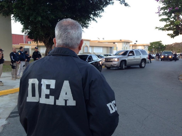 A DEA agent stands facing other agents and vehicles in a parking lot.