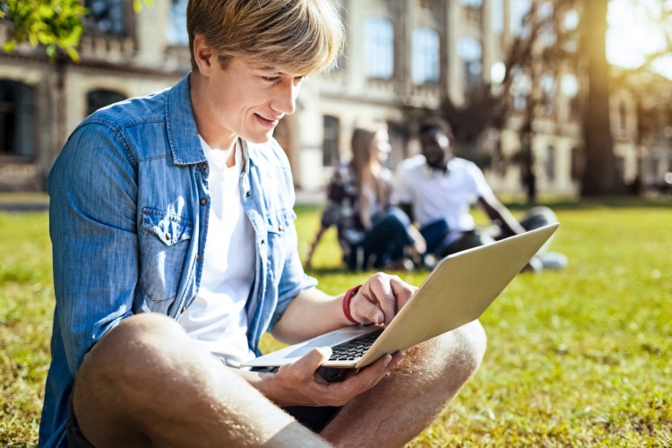 A student sits on a sunny lawn in front of a college building studying on a laptop.