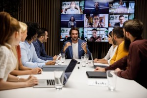 A manager leads a meeting with colleagues at a conference table and on a video screen.
