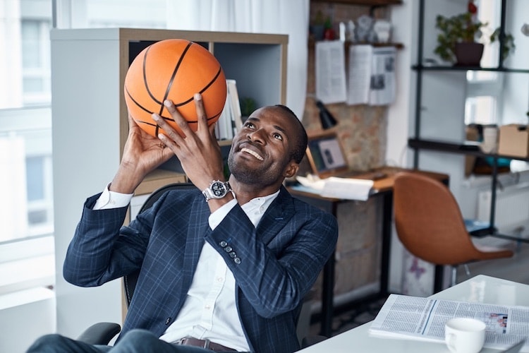 A businessman holds up a basketball while sitting at a desk in an office