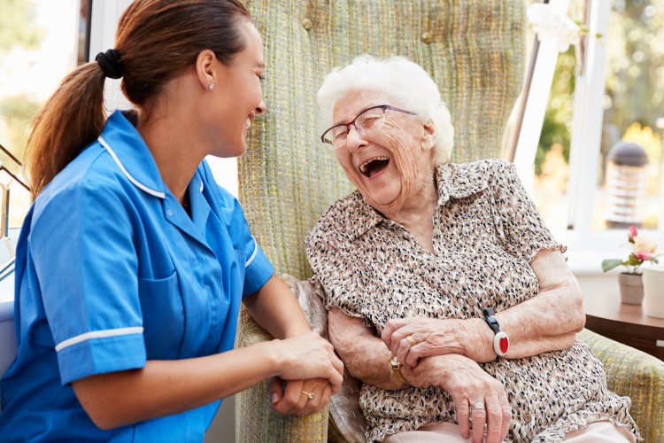 A young gerontology nurse in blue scrubs is seated beside a laughing elderly person