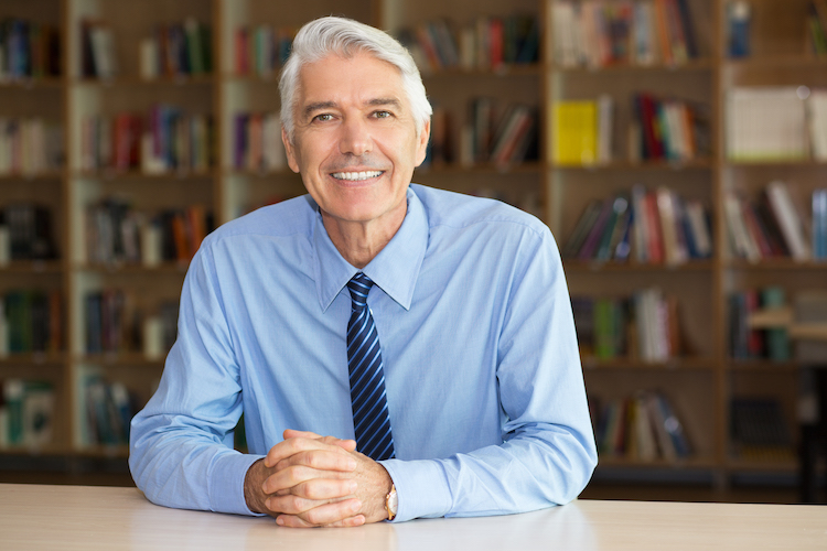 Portrait of senior Caucasian businessman or teacher wearing shirt and tie sitting at workplace in library, looking at camera and smiling