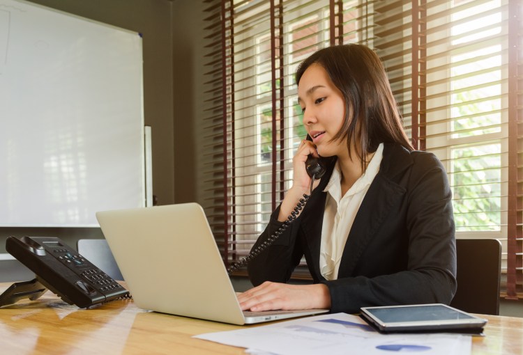 A hospital administrator works on a laptop while on the phone at her desk