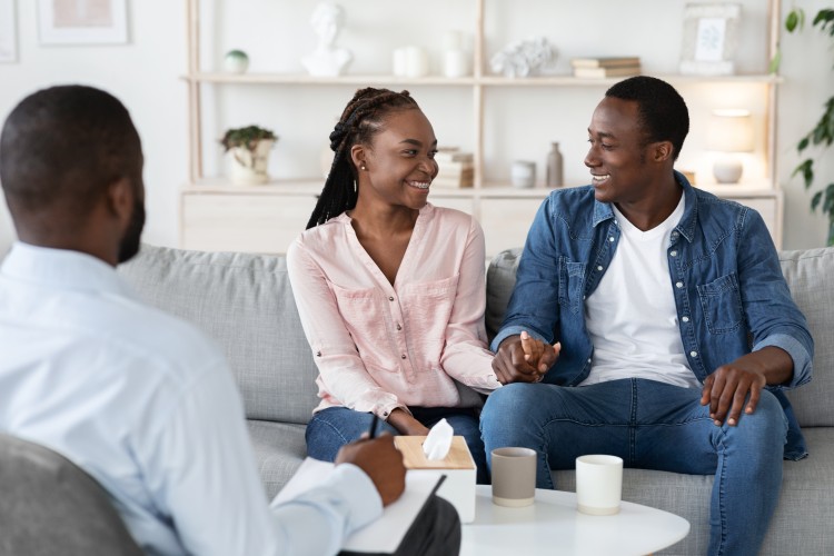 A young couple is smiling and holding hands during a session with a marriage and family therapist in an office.