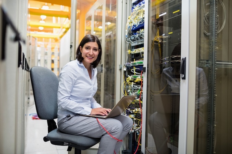A network architect works on her laptop in a server room.