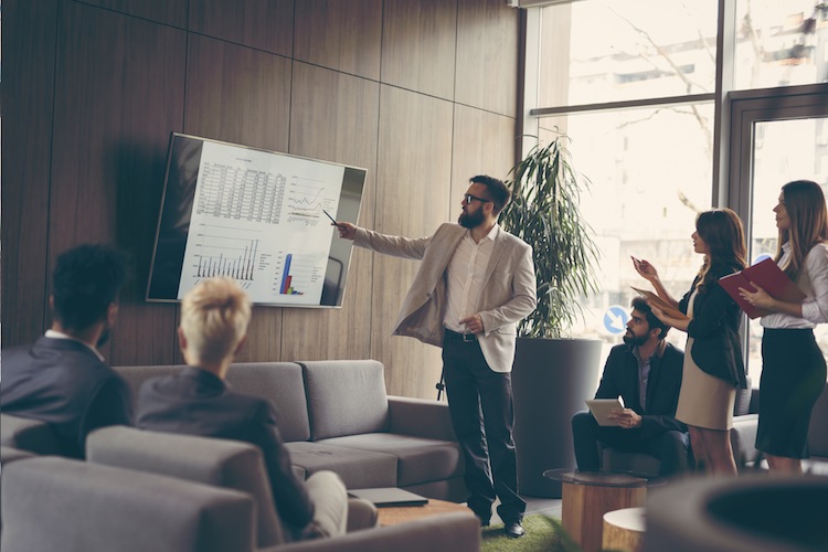 A business analyst points at a digital display while giving a presentation to a group of coworkers