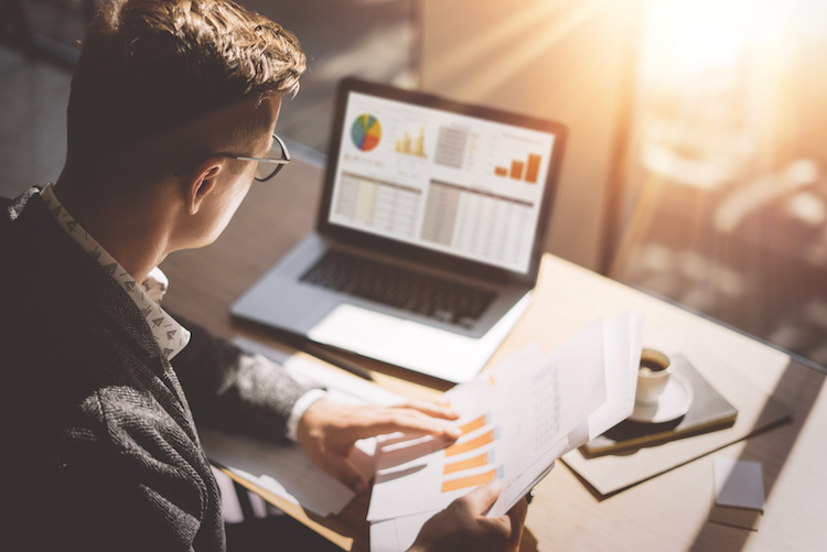 Young finance market analyst in eyeglasses working at sunny office on laptop while sitting at wooden table. Businessman analyze document in his hands. Graphs and diagram on notebook screen. Blurred