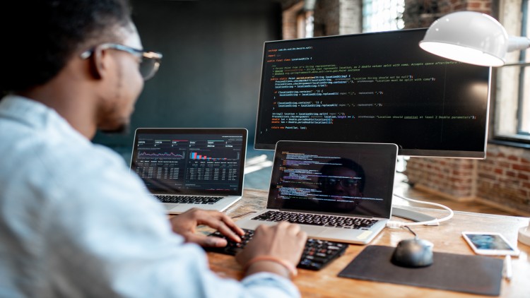 A software developer is seated at a desk in front of three computer monitors. 