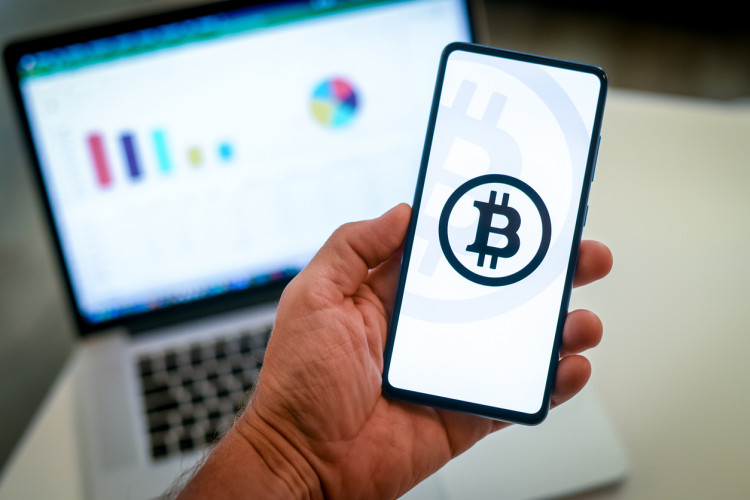A hand holding a smartphone displaying the Bitcoin logo.