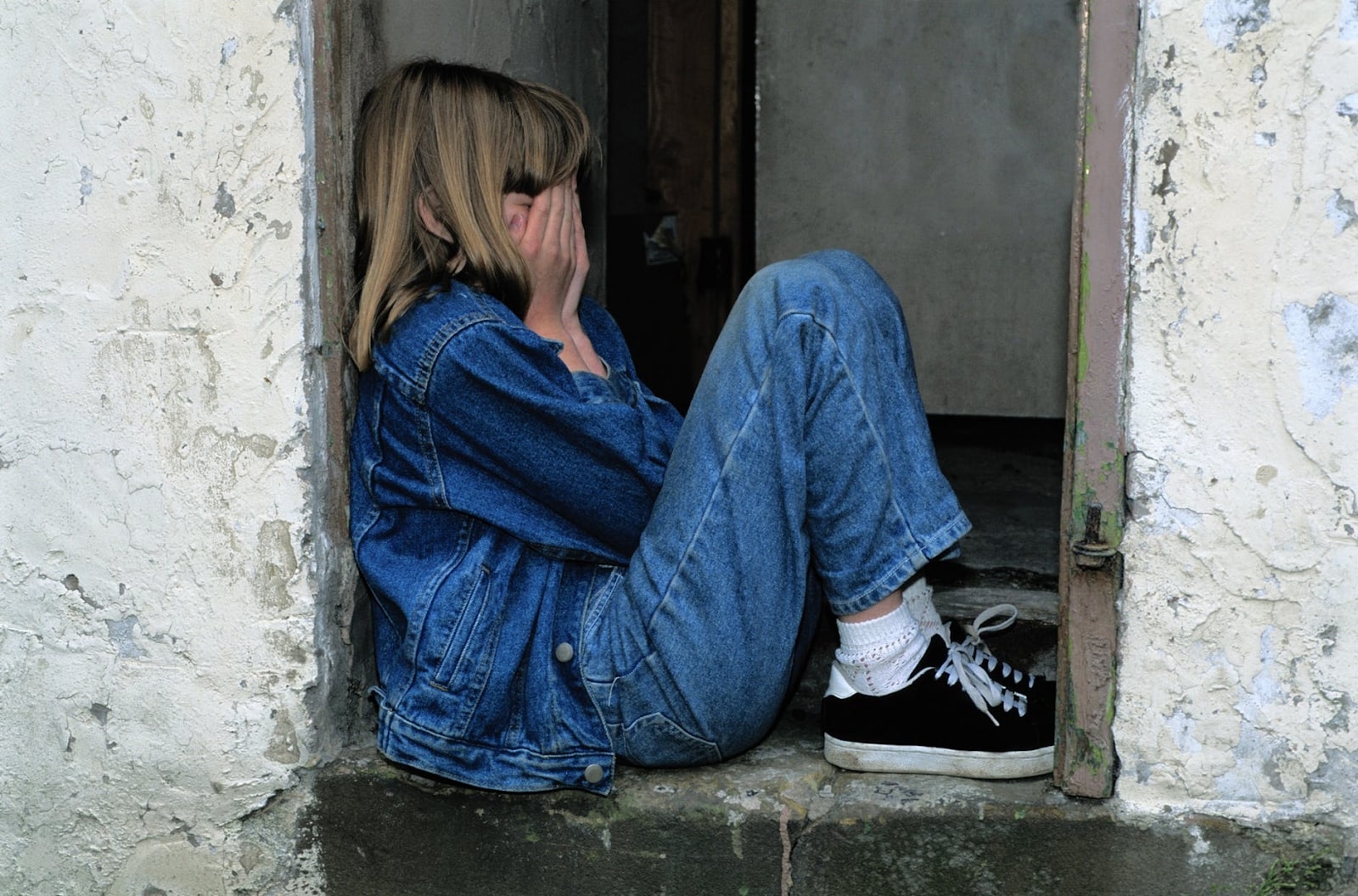 A concept photo of loneliness, showing a young girl from the side, wearing denim and covering her face, sitting in a small alcove.