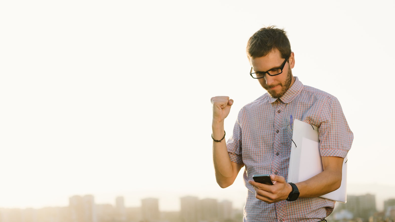 Working professional carrying documents outdoors pumps his fist while looking at a smartphone